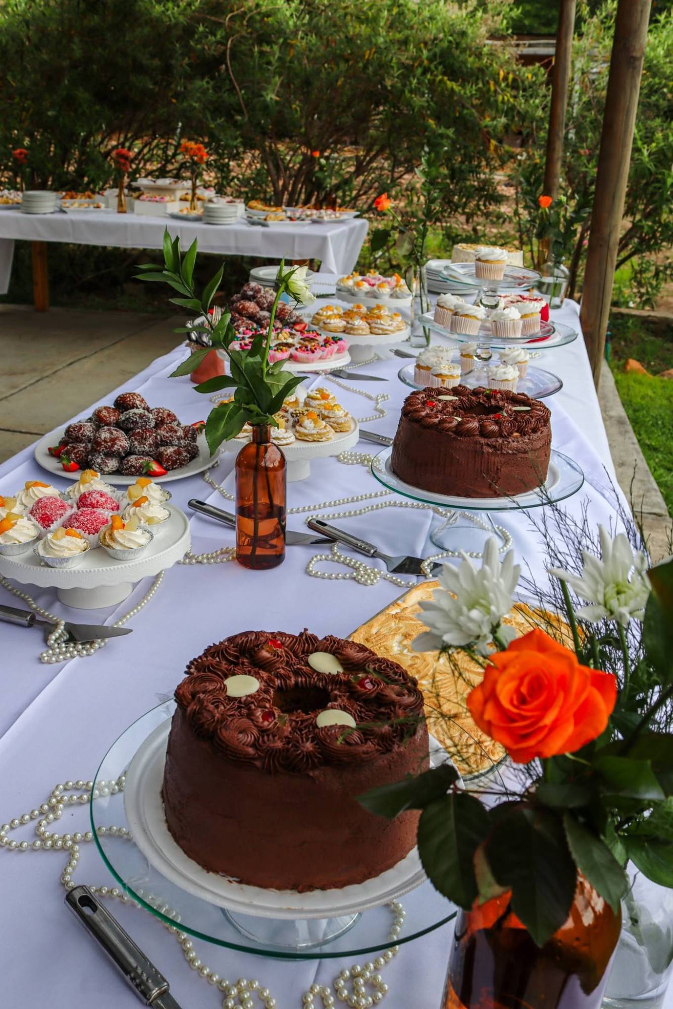 Dessert table by Creations catering: chocolate ring cakes on glass cake stands, cupcakes, coconut balls, cream puffs and a meringue tart on a white-clothed table dressed with pearl strands and orange roses in amber bottles