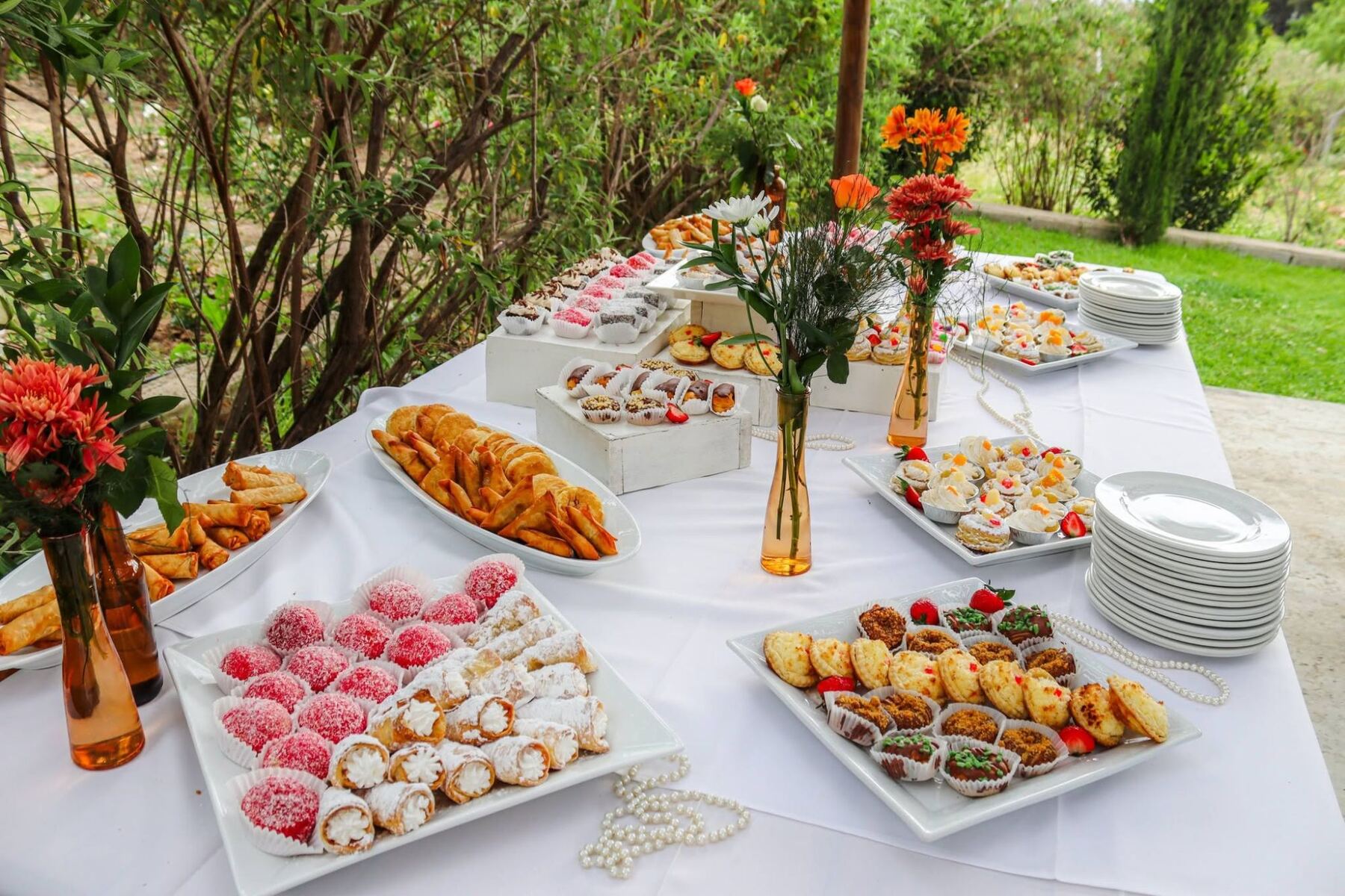 Catering spread by Creations: platters of pink coconut balls, cream-filled cannoli, spring rolls, samoosas, pastries and mini cakes on white crates, with stacked plates and orange blooms in amber glass vases in a garden