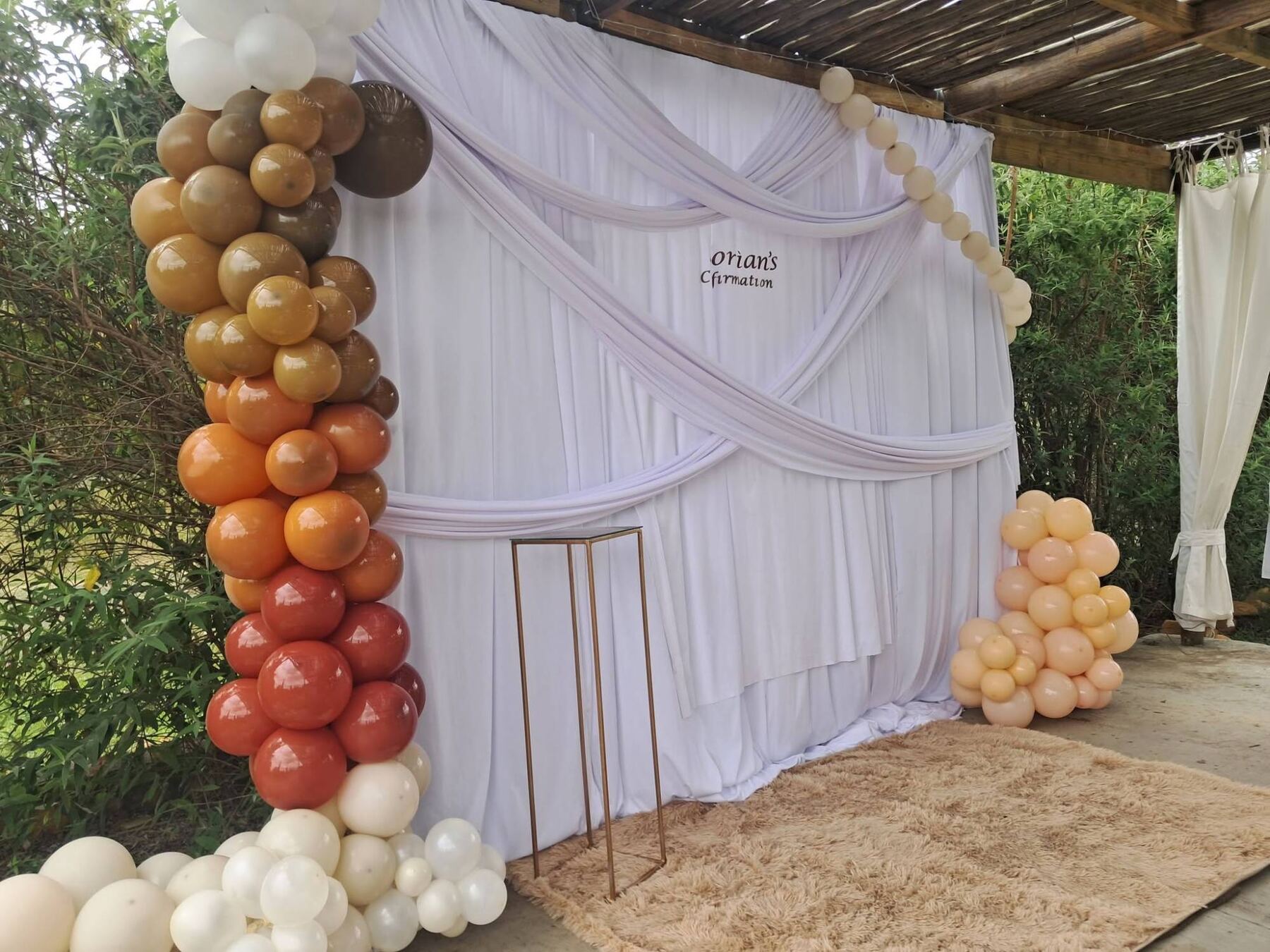 A confirmation photo backdrop by Creations: white draped fabric with a personalised name sign, an organic balloon garland in caramel, terracotta, rust and cream, a gold plinth and a fluffy beige rug under a thatched pergola