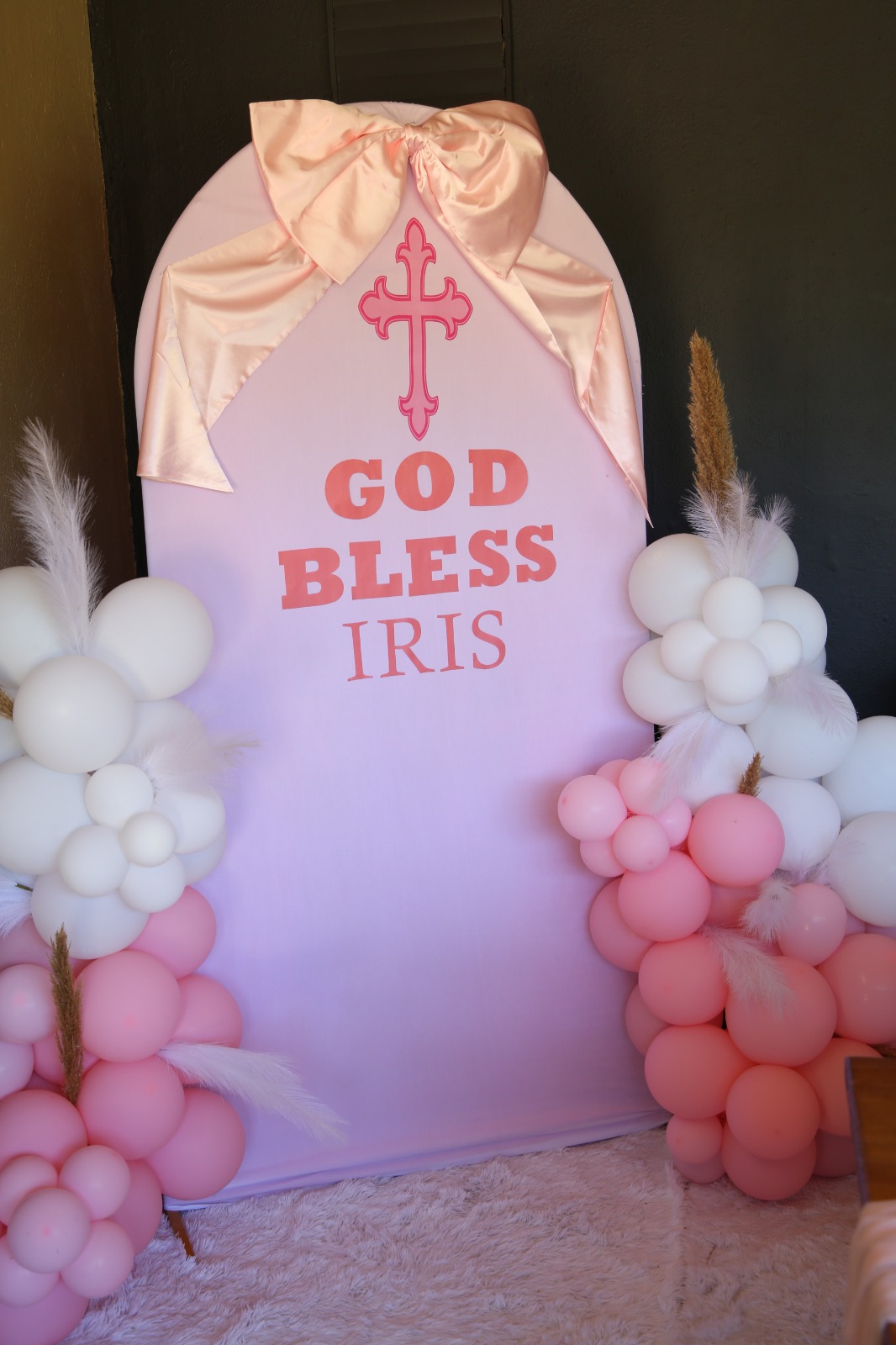A confirmation arch backdrop by Creations: a pale pink arch with a satin bow, a pink cross and a personalised blessing, flanked by white and pink balloon clusters with pampas feathers on a fluffy rug