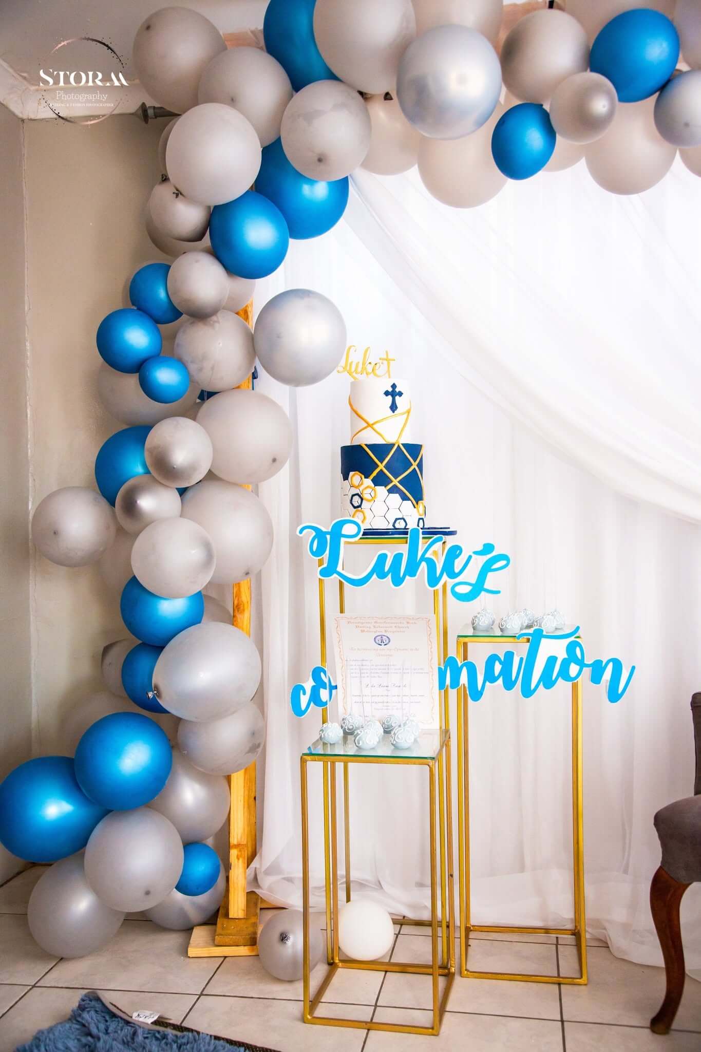 Confirmation decor by Creations: a blue and silver balloon garland over white draping, a two-tier navy and gold cross cake on gold plinths and a personalised name sign