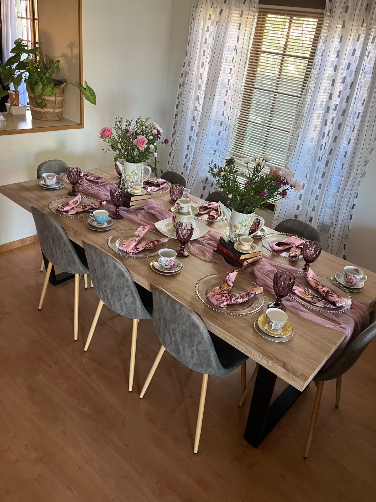 A vintage tea-party table by Creations: a wooden dining table with a dusty-pink tulle runner, glass-beaded chargers, floral napkins, mismatched china teacups, plum goblets, stacked books and jugs of fresh flowers