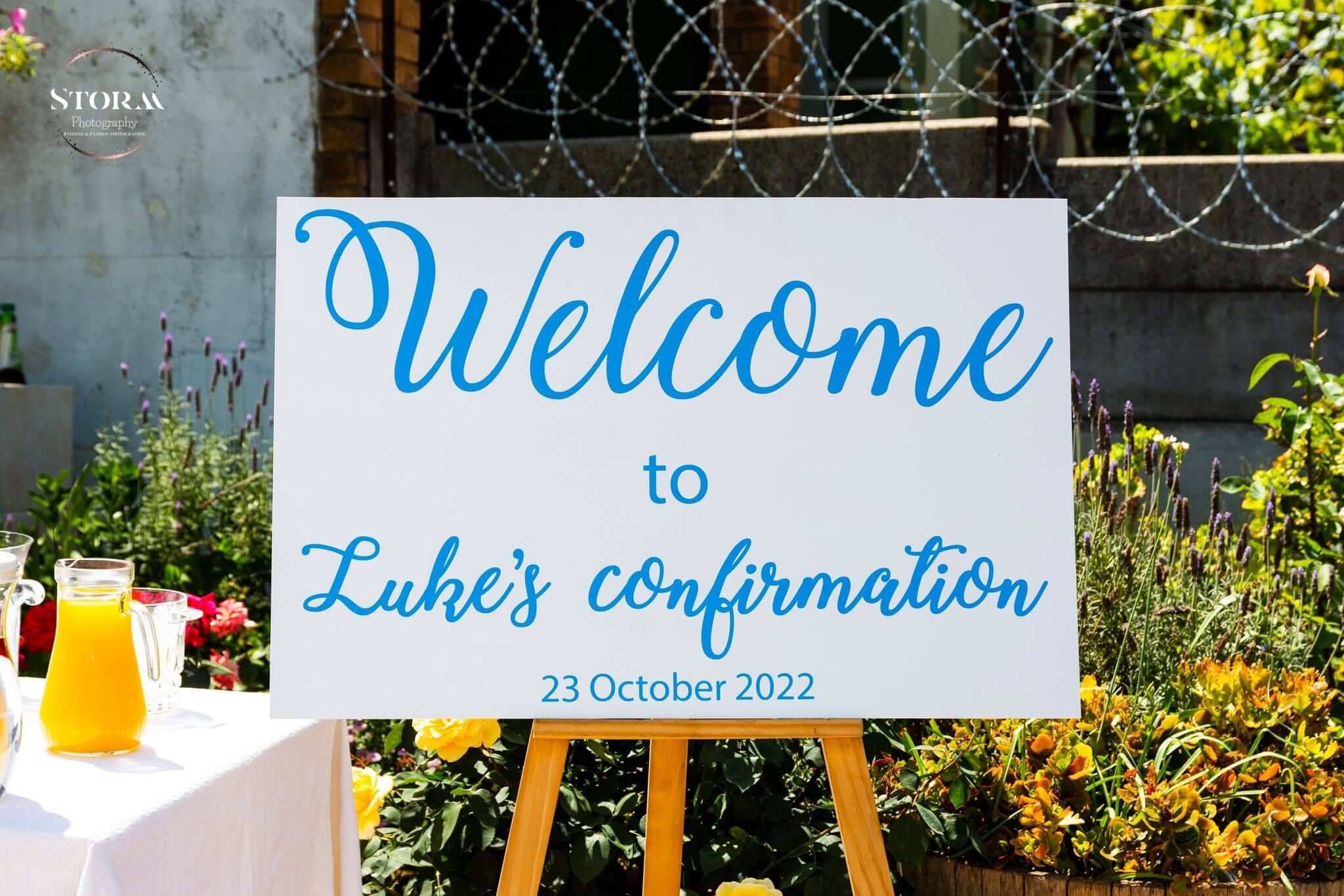 Confirmation decor by Creations: a hand-lettered blue welcome sign on a wooden easel among lavender and roses, with juice jugs on a white-clothed table