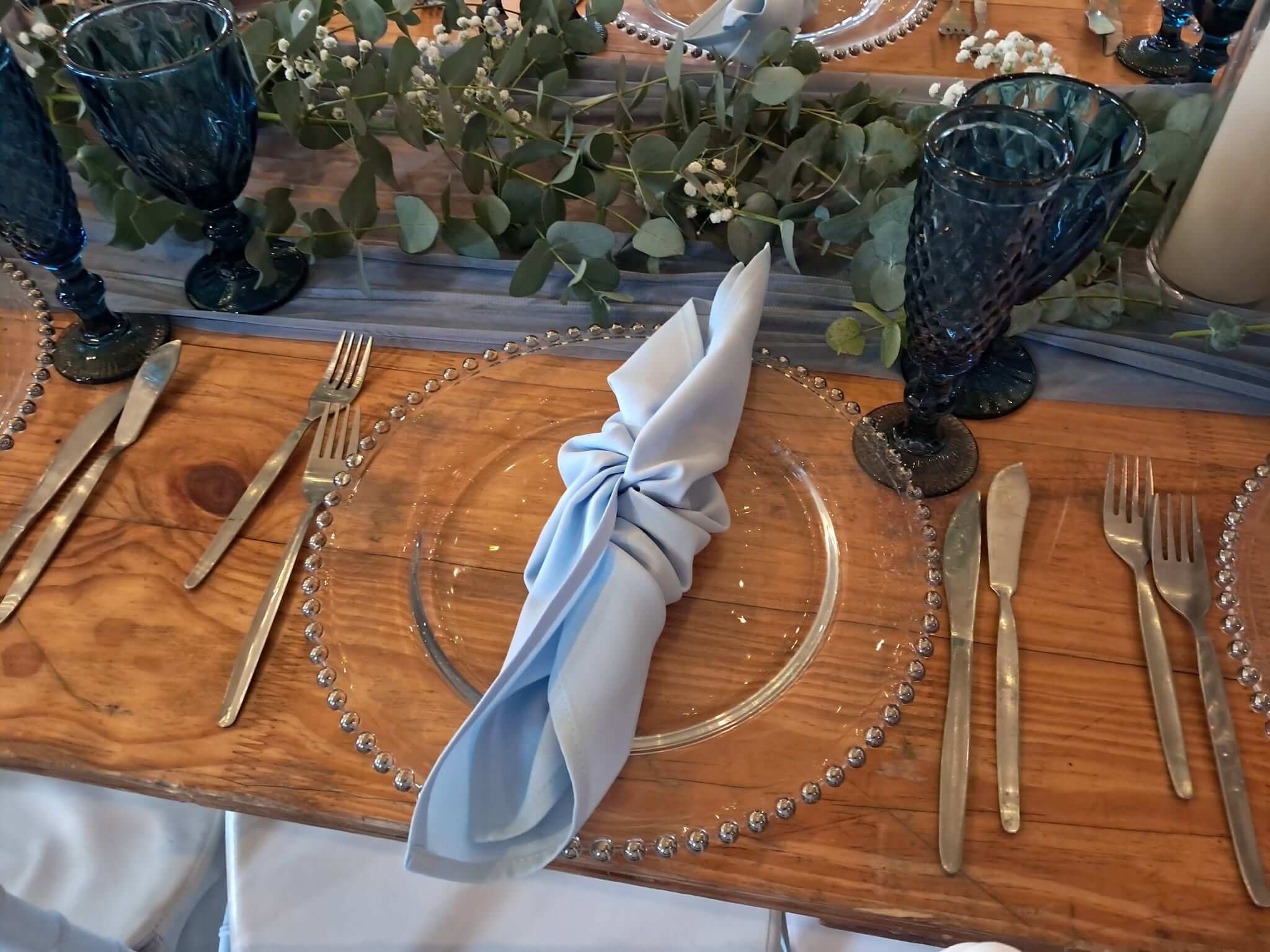 Wedding decor by Creations: a dusty blue napkin on a beaded glass charger plate, blue crystal goblets and a eucalyptus runner on a wooden table