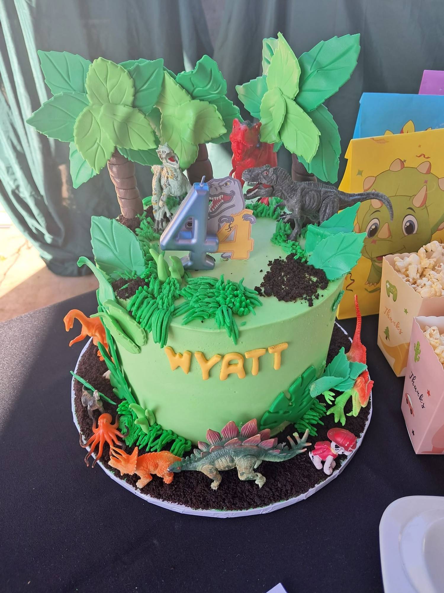 A dinosaur-themed birthday cake with green icing, sugar palm trees, chocolate crumb soil, toy dinosaurs and a number candle, beside dinosaur treat bags and popcorn boxes