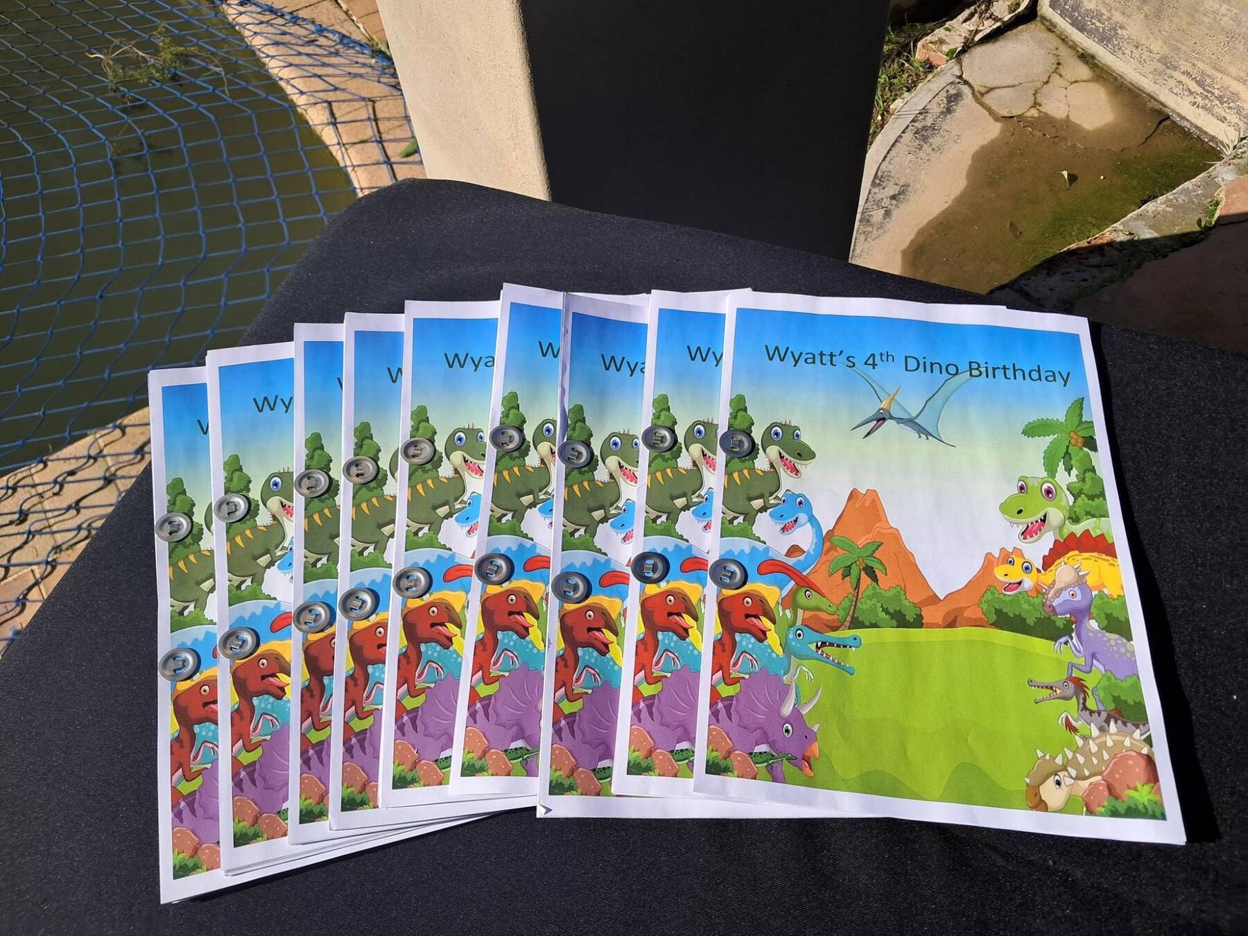 A fan of printed dinosaur-themed party booklets with a colourful prehistoric illustration, laid out on a black chair