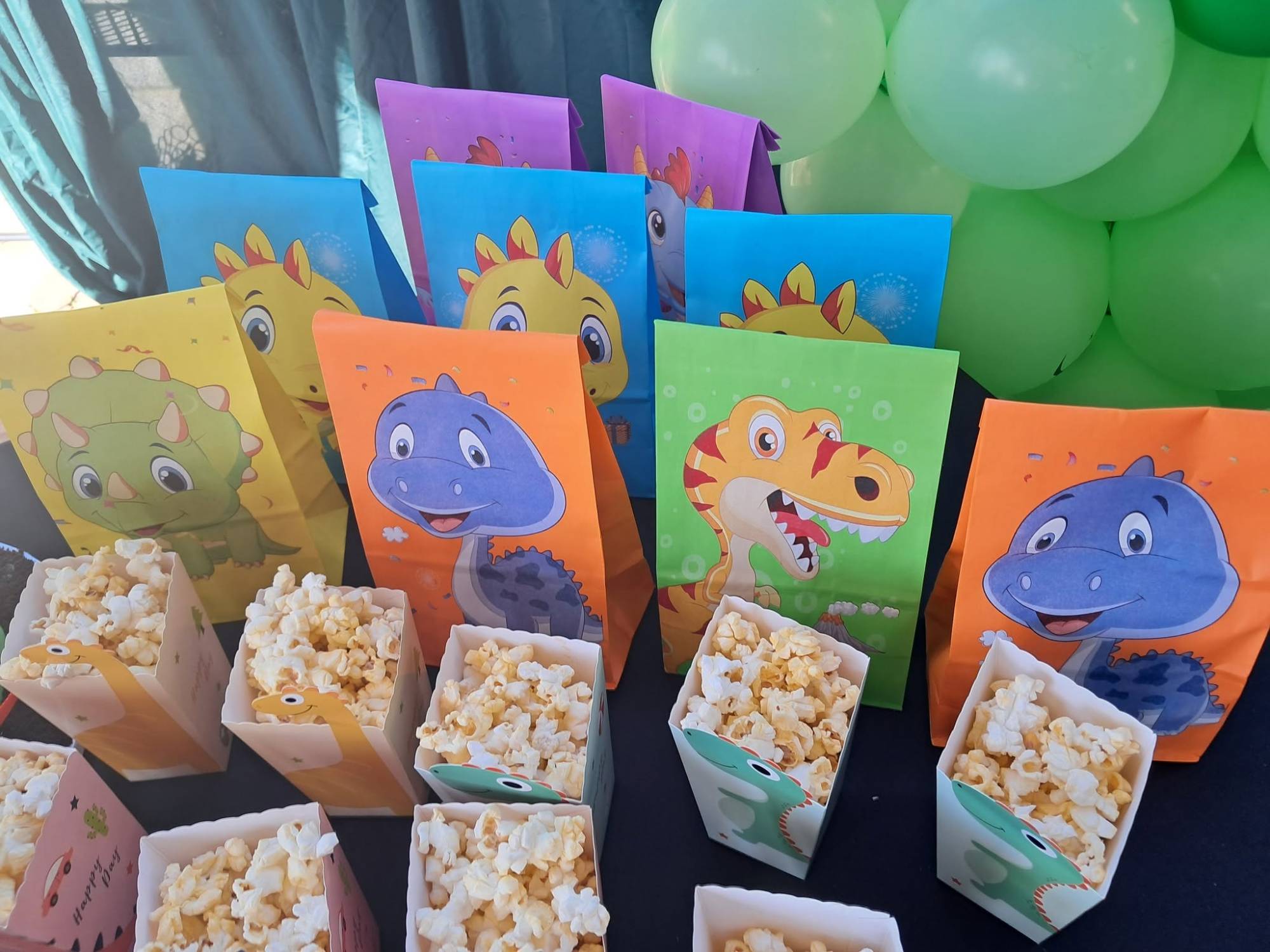 Colourful dinosaur treat bags in yellow, blue, orange, green and purple with popcorn boxes in front of a green balloon garland and teal drapes