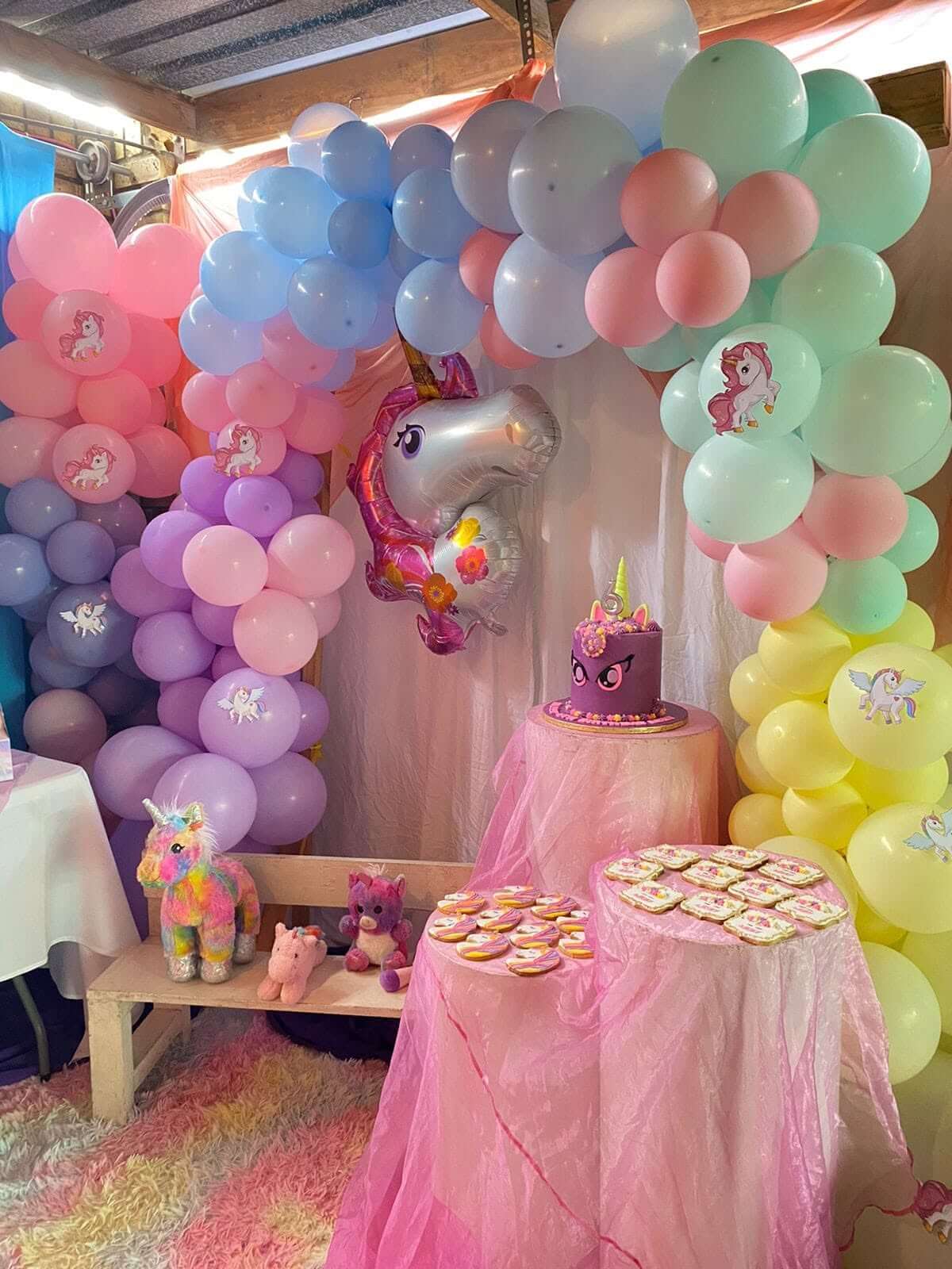 Unicorn kiddies party decor by Creations: a pastel rainbow balloon arch with unicorn balloons, a purple unicorn cake on a pink draped table and unicorn cookies
