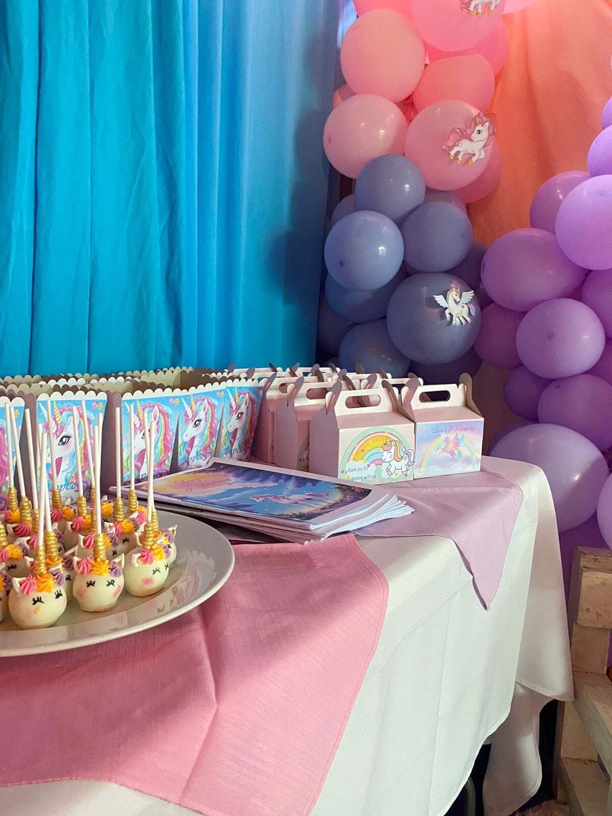 Unicorn party treats by Creations: unicorn cake pops, themed party boxes and popcorn holders on a pink and white table against a blue drape
