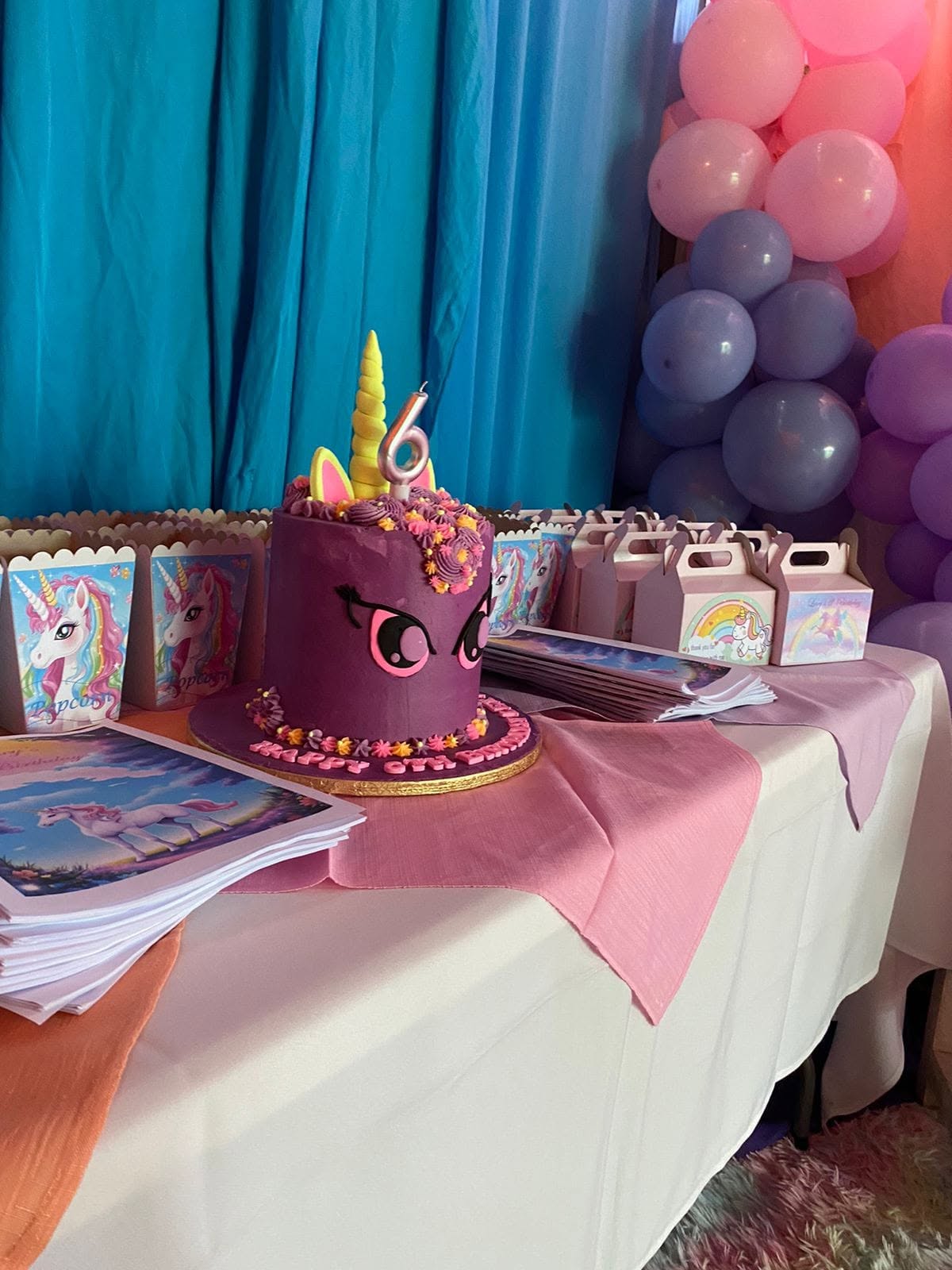A purple unicorn cake with a yellow horn, pink ears and a number candle on a white table with pink runners, unicorn popcorn boxes and treat boxes, in front of blue drapes and a pink, lilac and purple balloon garland