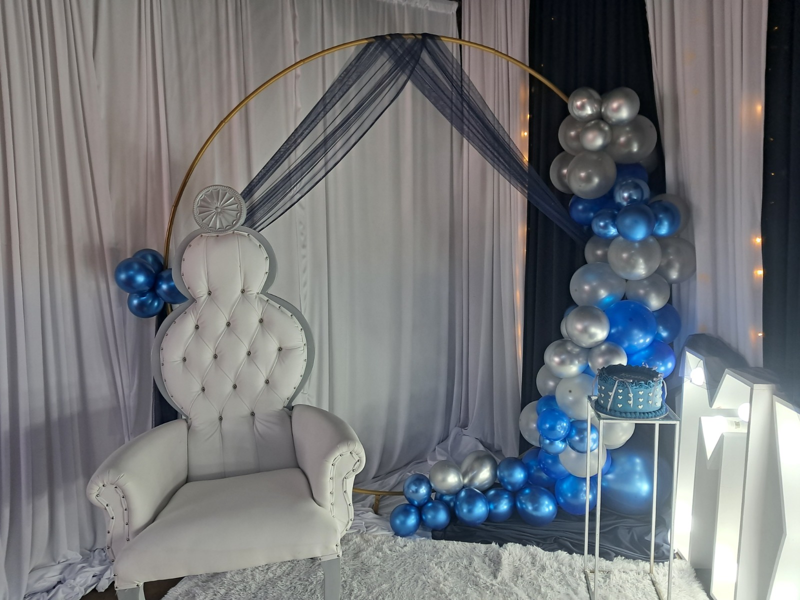 Matric photo backdrop by Creations: a white throne chair beside a gold ring arch draped with navy tulle, a blue and silver balloon garland, a navy cake and light-up marquee letters
