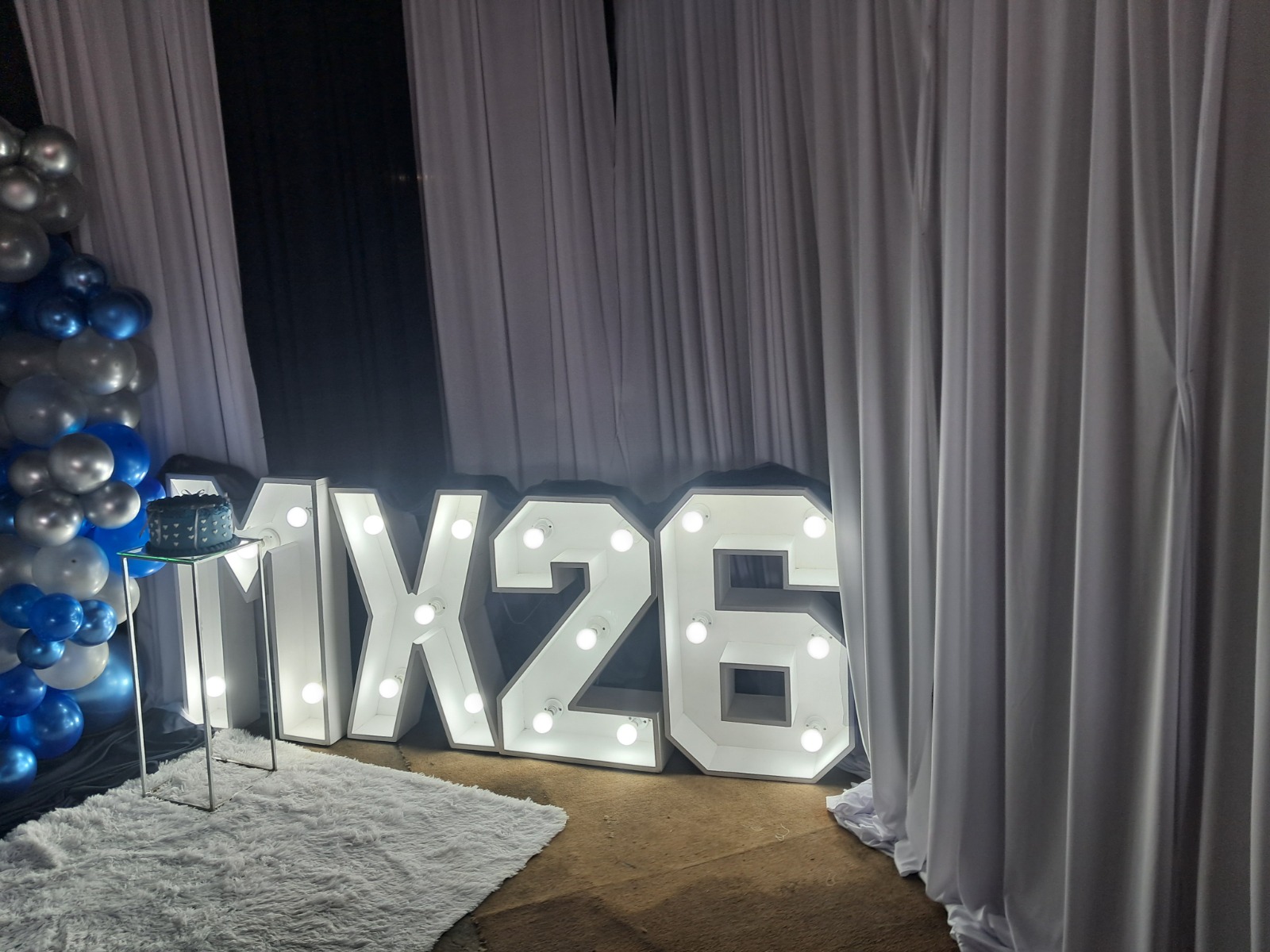 Matric celebration decor by Creations: white light-up marquee letters and numbers beside a cake stand and a blue and silver balloon column, in front of white and navy draping