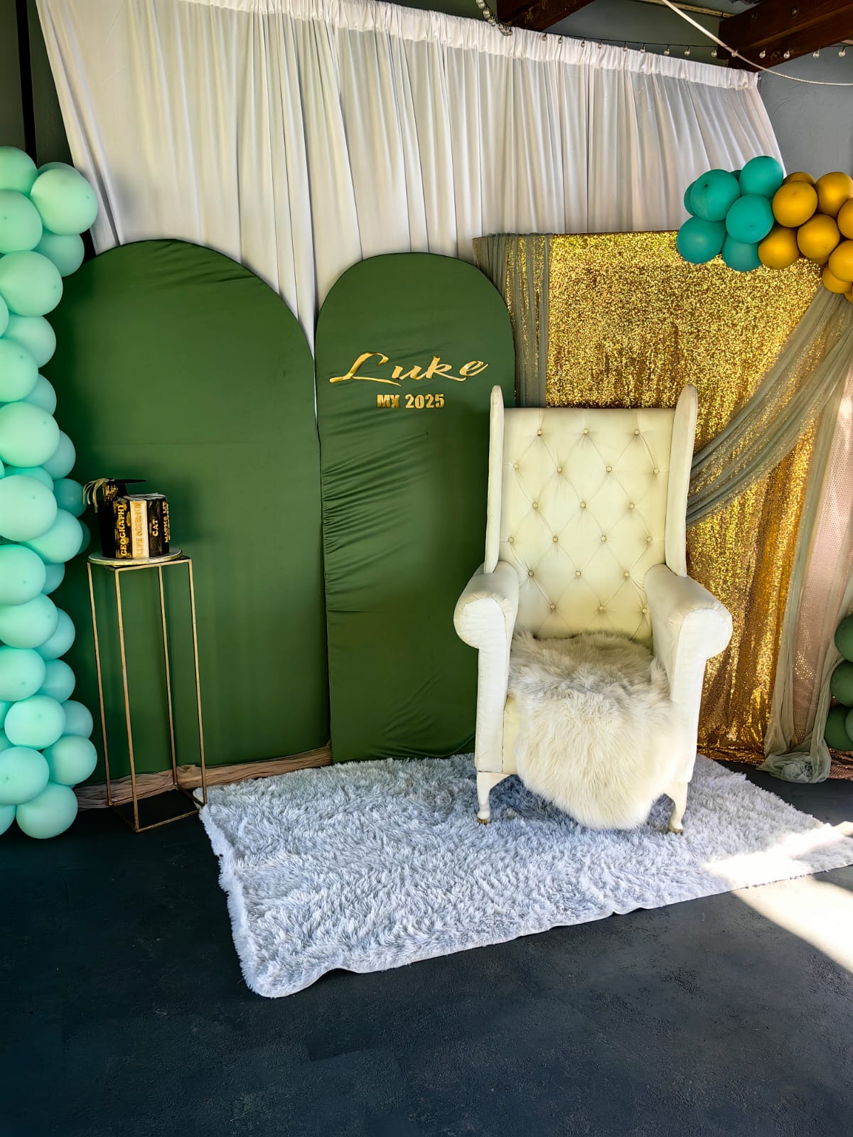 A matric photo corner by Creations: an emerald green arched backdrop with a gold name sign, a white wingback chair on a fluffy rug, a gold sequin panel with draping, mint and gold balloon clusters and a graduation cake on a gold plinth