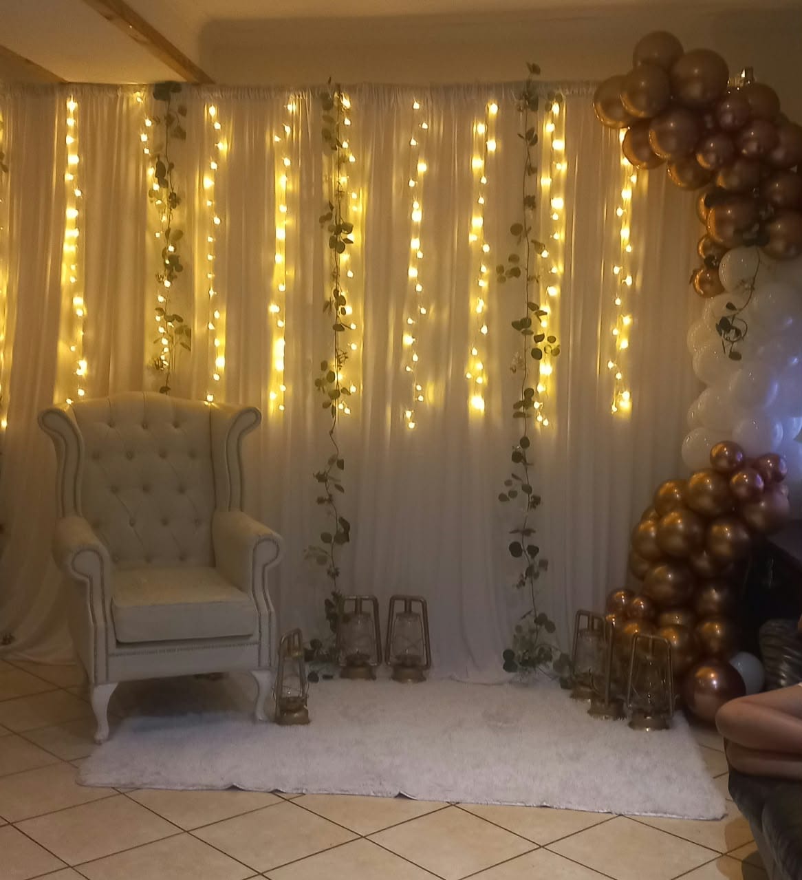 Photo backdrop by Creations: a white wingback chair before white draping hung with warm fairy lights and trailing greenery, with brass lanterns and a gold, white and rose-gold balloon garland