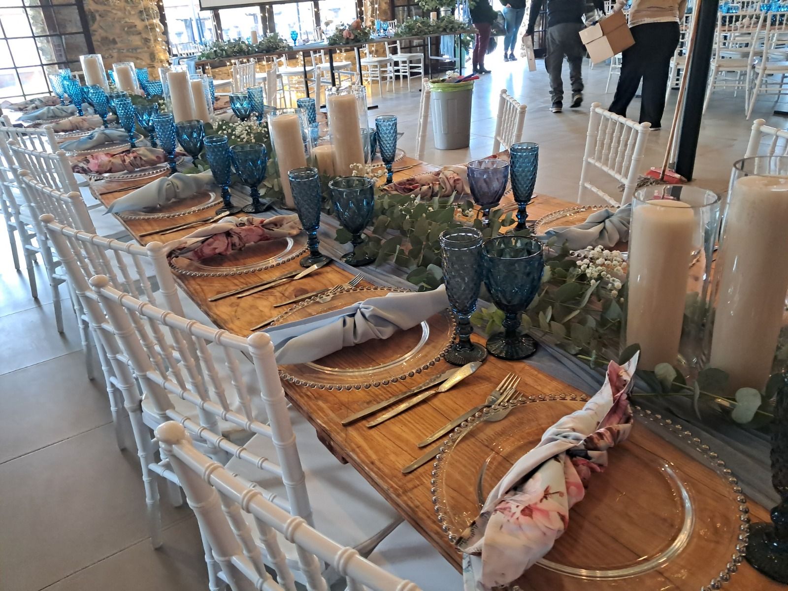 Wedding decor by Creations: a long wooden banquet table set with blue crystal goblets, beaded charger plates, dusty-blue and floral napkins, pillar candles and a eucalyptus runner, with white Chiavari chairs
