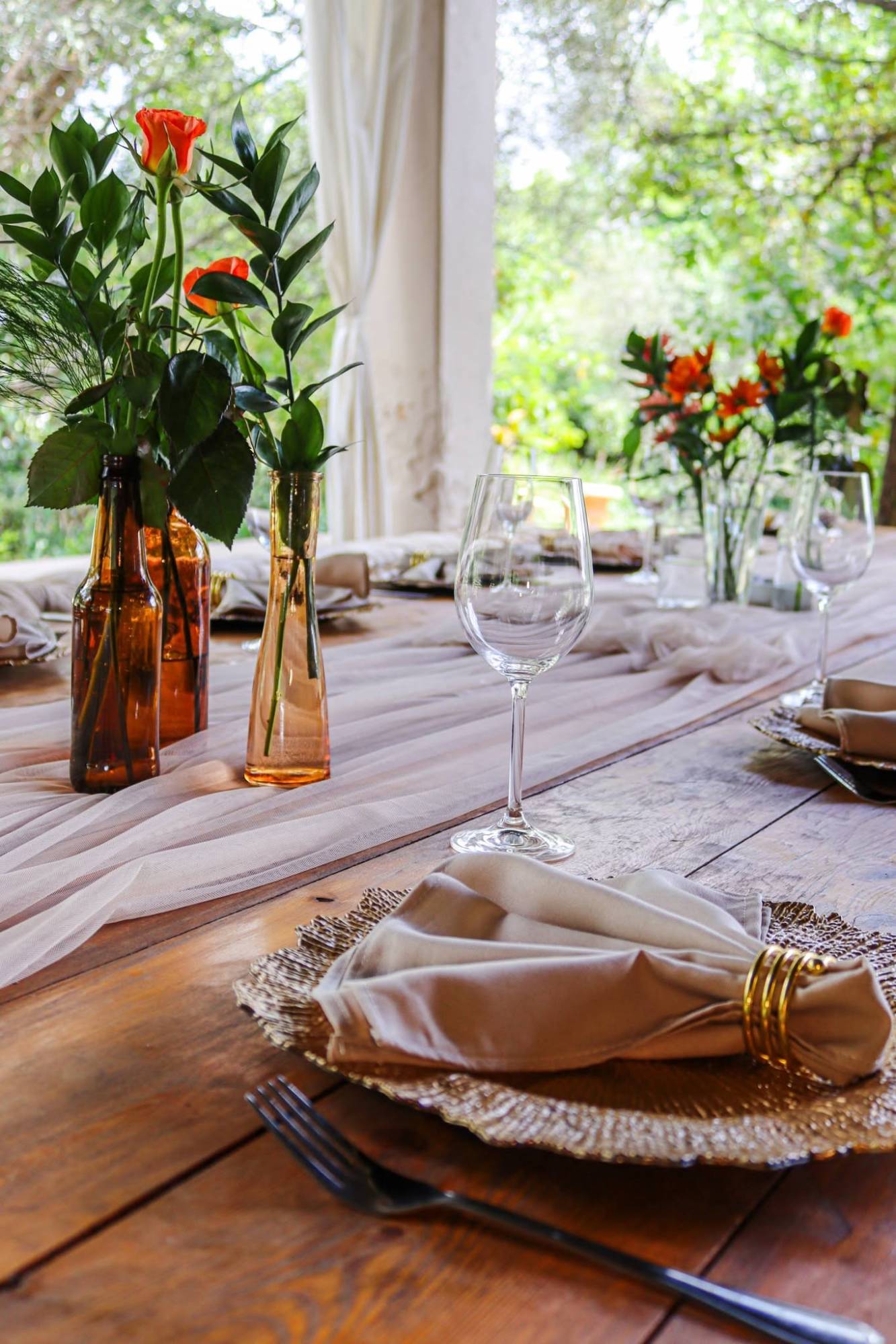 Amber bottle vases with orange roses and pink gerberas on a blush tulle runner, set beside a wine glass and a gold charger plate with a napkin and gold ring
