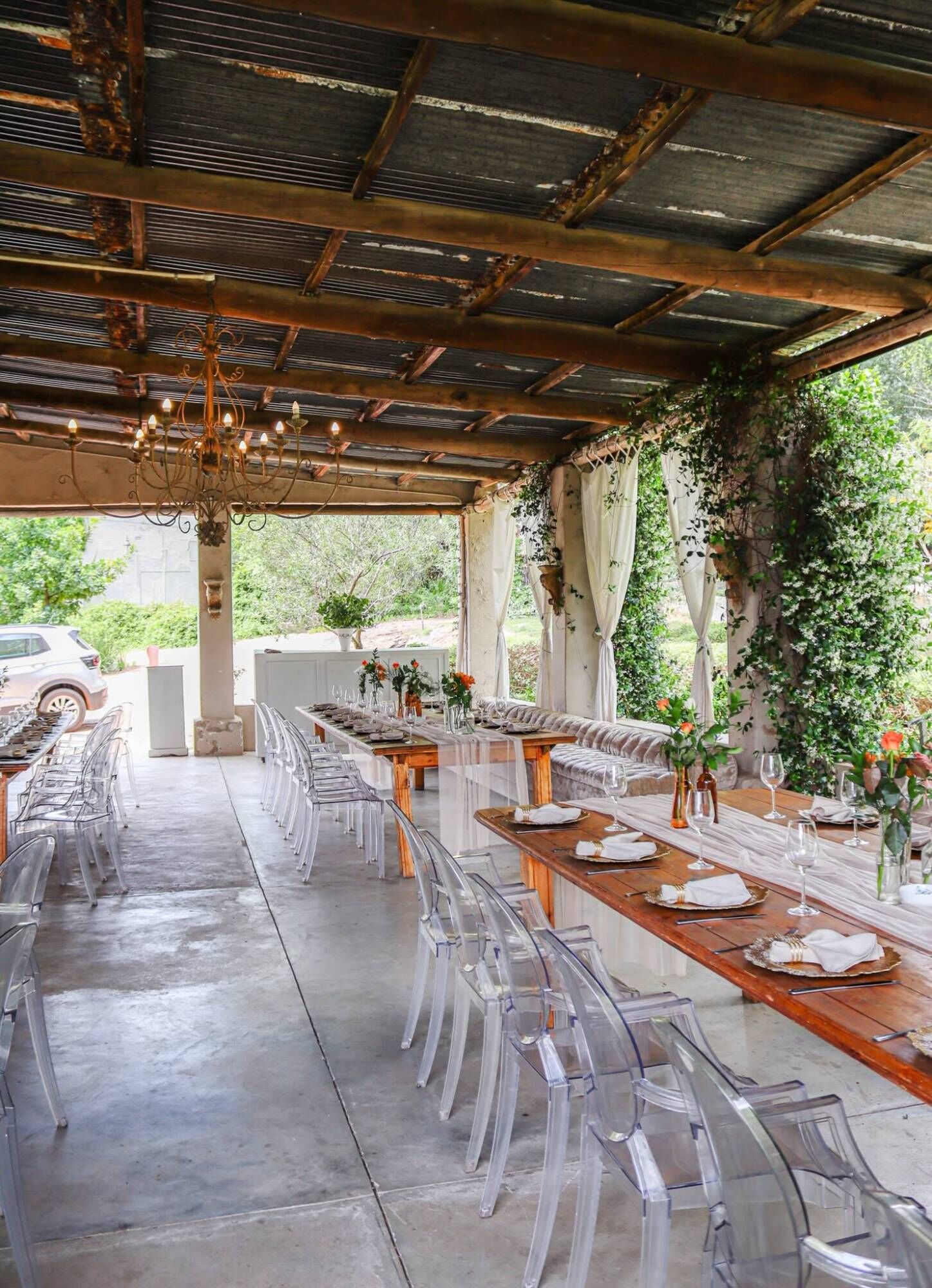 A garden pavilion under a rusted tin roof with a chandelier, long rustic farm tables with gold chargers and clear ghost chairs, and white curtains with climbing jasmine