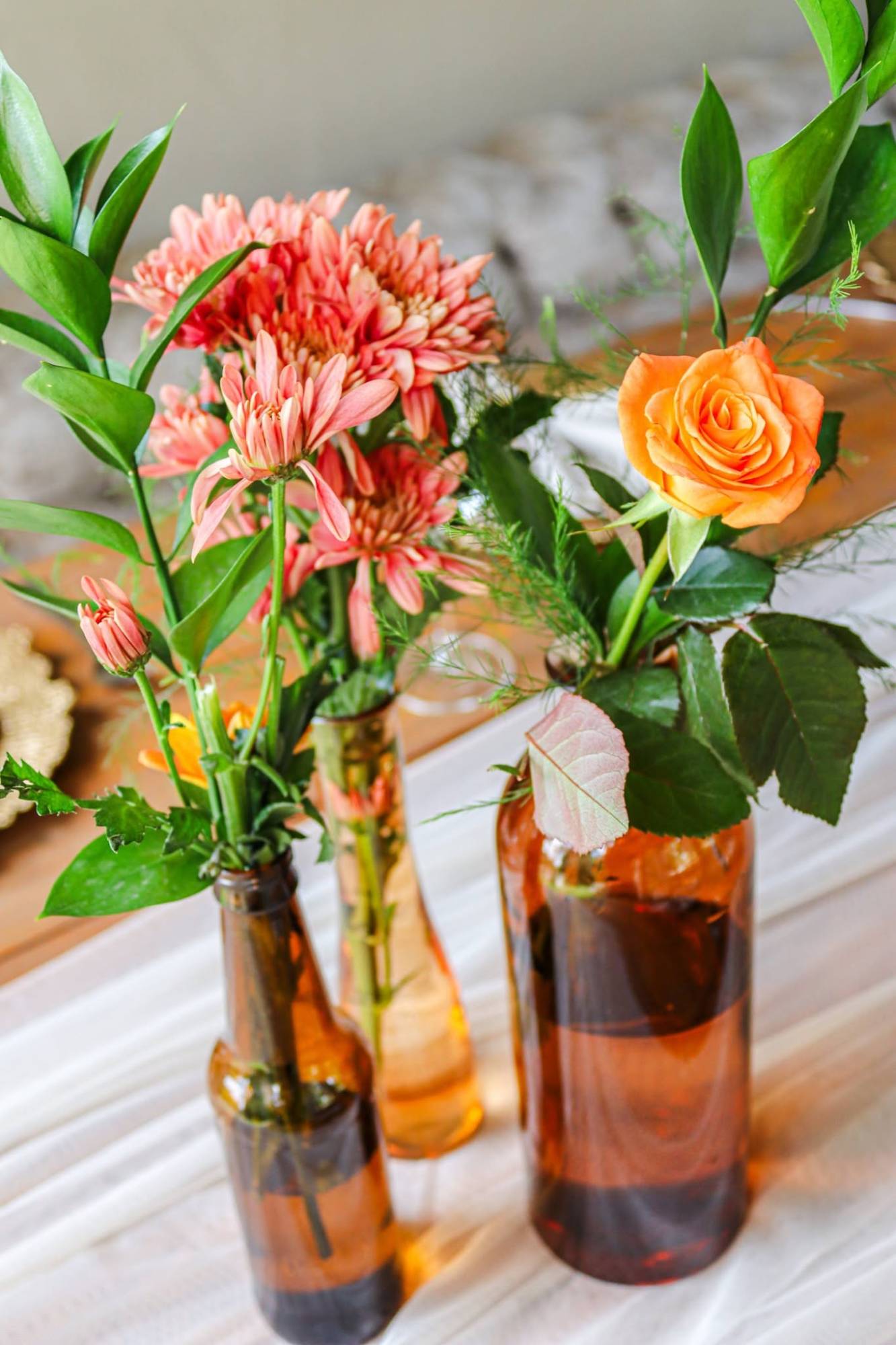 Close-up of amber glass bottle vases holding pink chrysanthemums, an orange rose and green foliage on a white runner