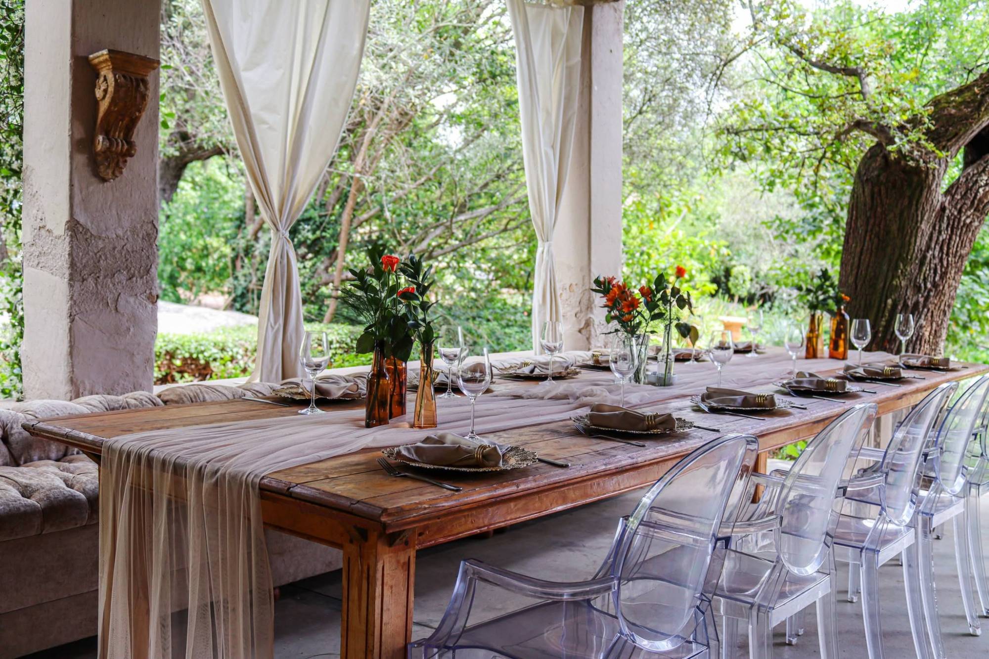 A long rustic farm table dressed with a blush tulle runner, gold charger plates and amber bottle vases, beside clear ghost chairs in a leafy garden pavilion