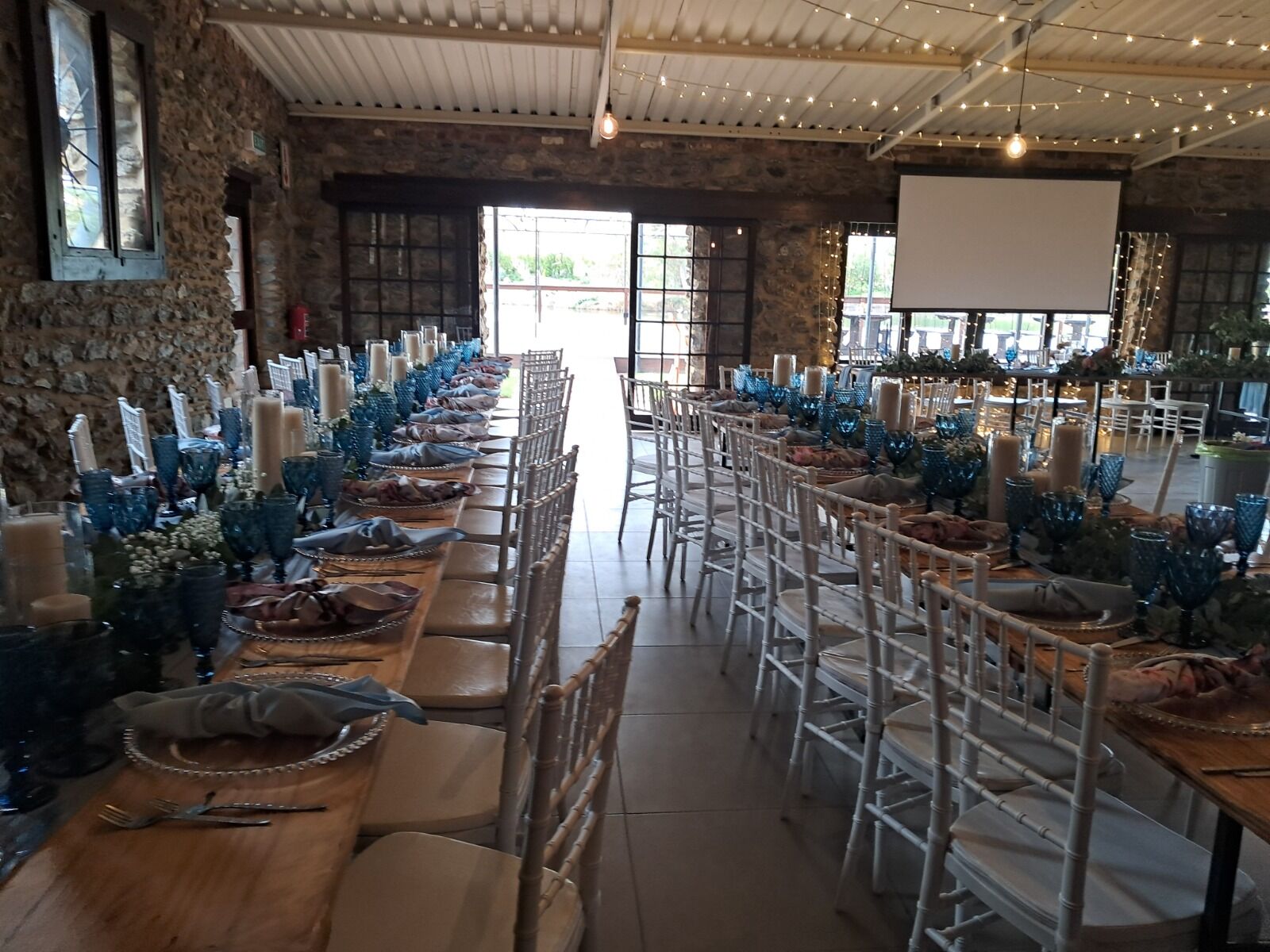 A stone-walled barn venue set with long wooden banquet tables, blue crystal goblets, pillar candles and white Chiavari chairs under festoon lighting