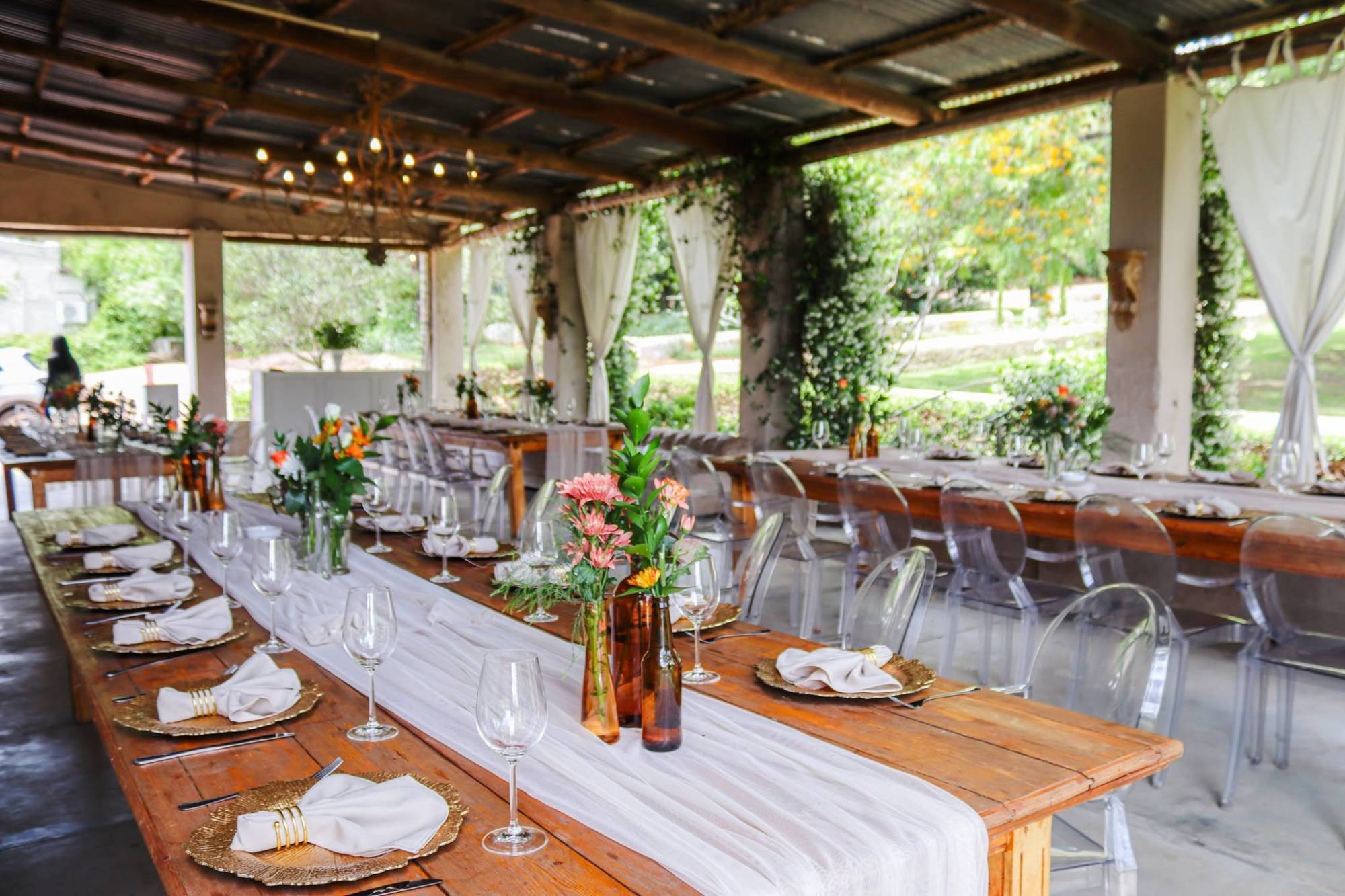 A covered garden pavilion set with long rustic farm tables, gold charger plates, amber bottle vases of orange blooms and clear ghost chairs under a chandelier