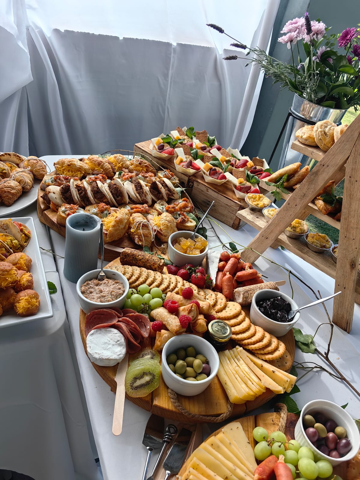Catering by Creations: a styled grazing table of cheese boards, canape boxes, sliders, croissants and pastries beside a wooden ladder stand, with a vase of fresh flowers