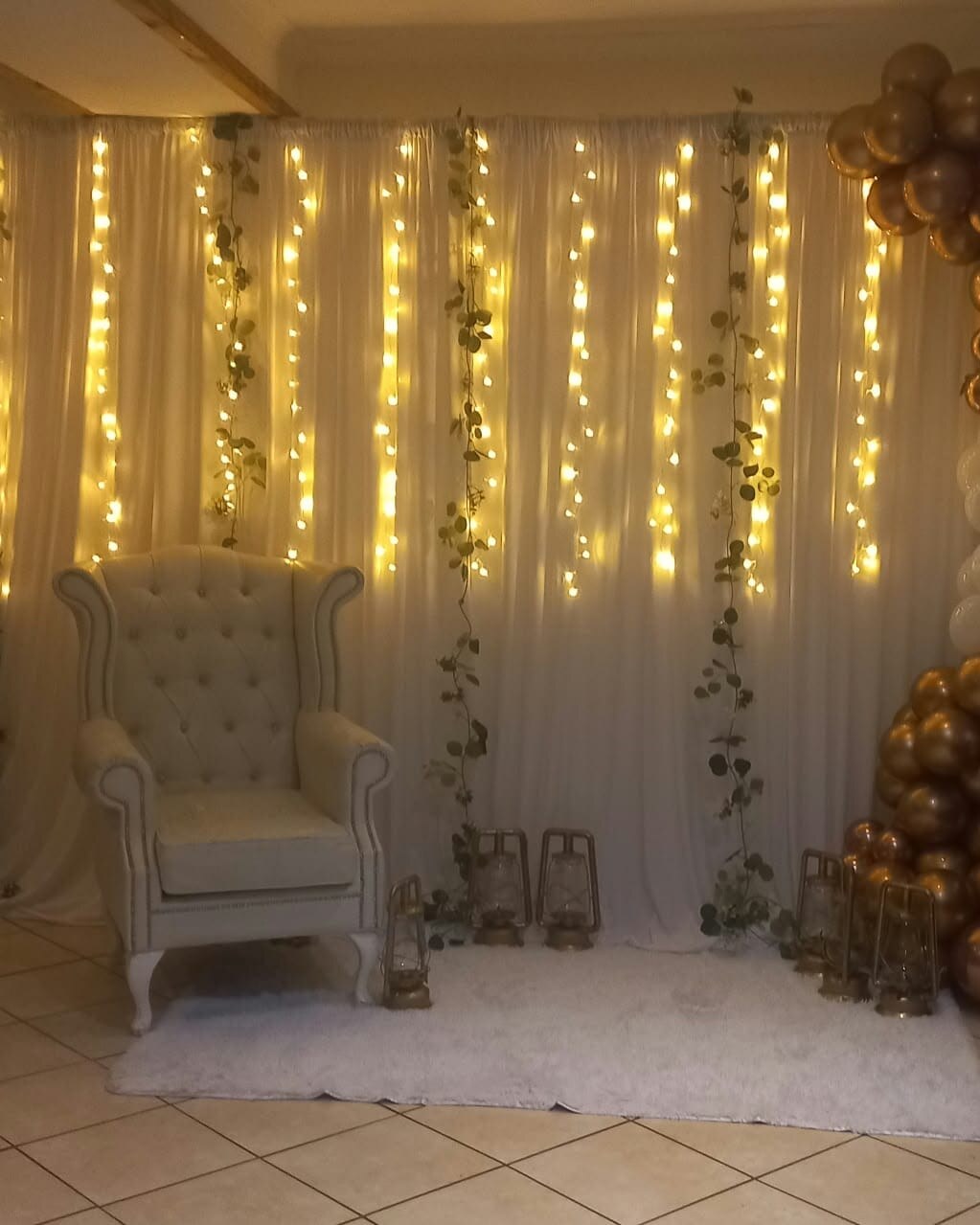 A glowing celebration photo backdrop with curtains of fairy lights, trailing eucalyptus, a white wingback chair and gold lanterns