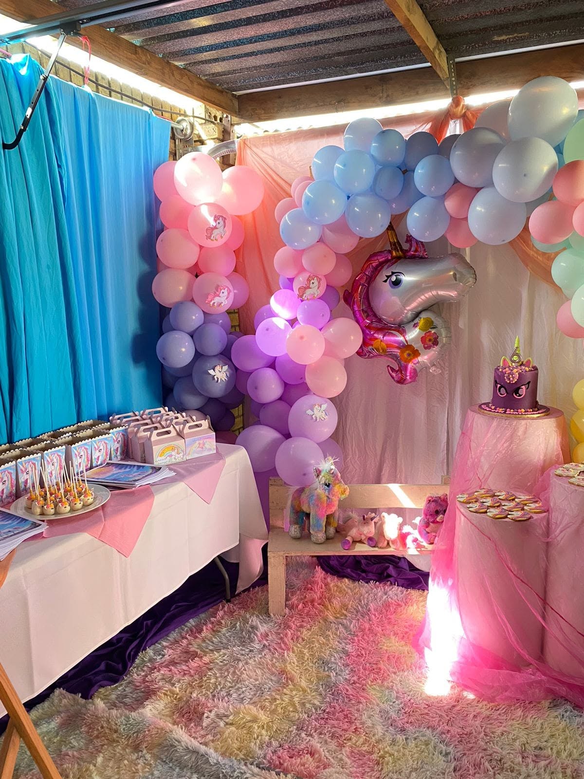 A unicorn-themed children's party corner with a pastel pink, blue and lilac balloon garland, a foil unicorn balloon, a plush toy bench and a dessert table with a purple unicorn cake