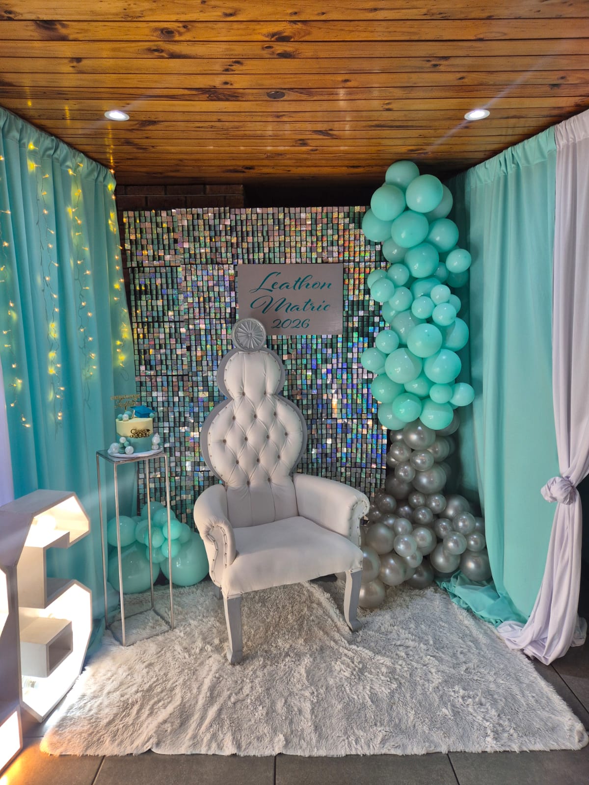 Matric photo backdrop by Creations: a high-backed white throne chair before a holographic sequin wall, mint drapes with fairy lights, a mint and silver balloon column, a personalised sign and a graduation cake