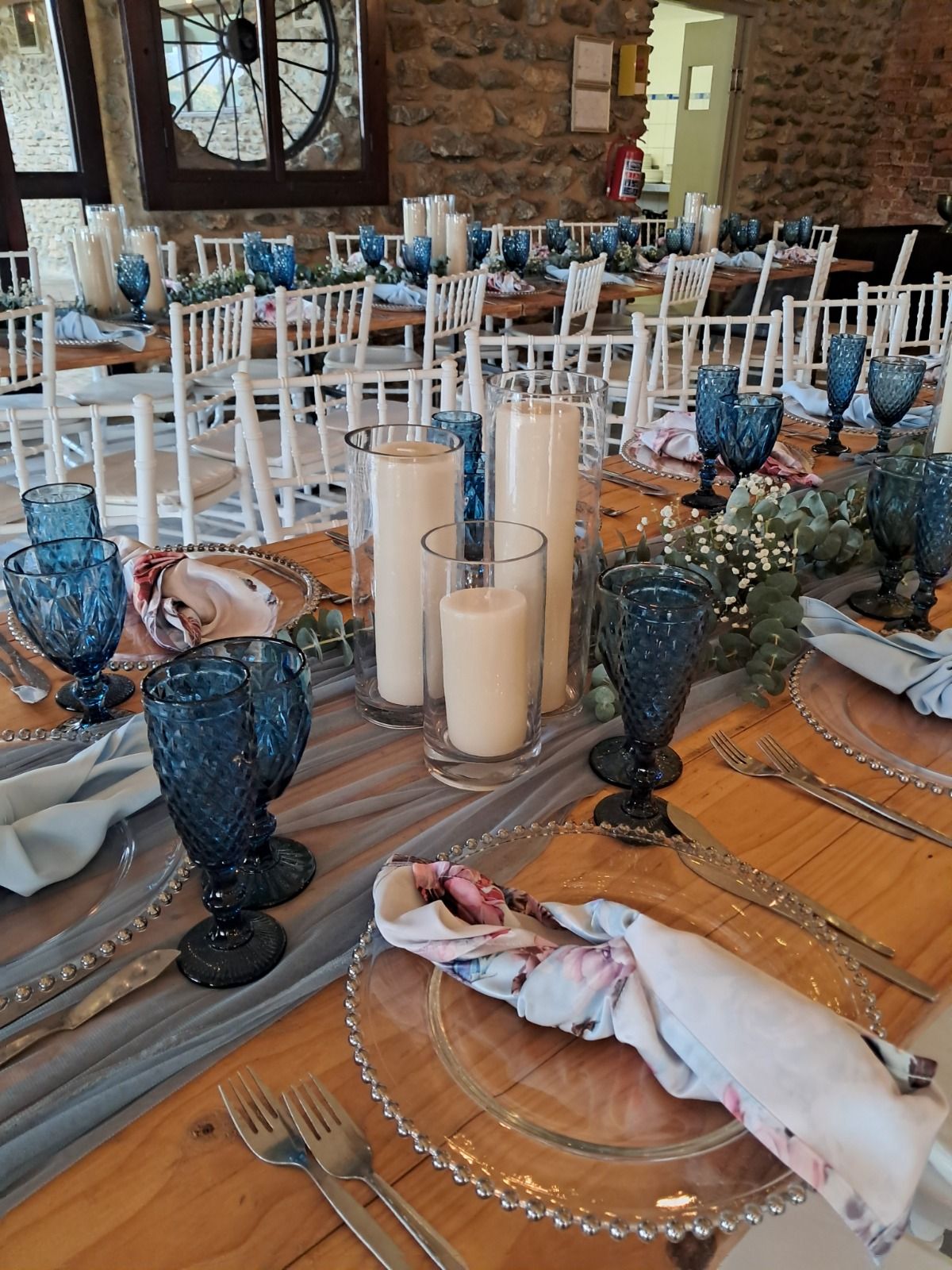 Wedding decor by Creations in a stone barn venue: blue crystal goblets, beaded charger plates, floral and dusty-blue napkins, pillar candles in glass hurricanes and a eucalyptus and gypsophila runner, with white Chiavari chairs